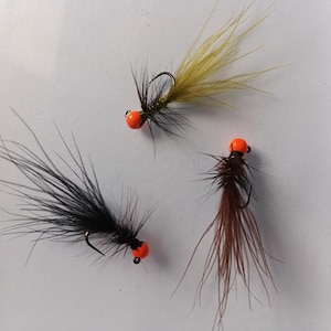 Può includere: Tre mosche da pesca con teste di perline arancioni e ami neri. Una mosca ha un corpo nero, una ha un corpo marrone e una ha un corpo giallo. Le mosche sono progettate per la pesca.