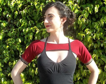 May include: A burgundy shrug with short sleeves, featuring black trim and a braided halter strap. The shrug is worn over a black top. The background is a lush green hedge.