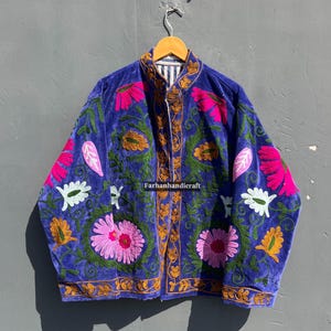 Puede incluir: Chaqueta de terciopelo azul con intrincados bordados florales en rosa, blanco y dorado. La chaqueta tiene un cuello alto y el texto "Farhanhandicraft". Cuelga de una percha de madera.