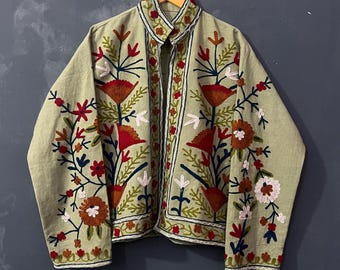 Op de afbeelding: Lichtgroene jas met gedetailleerde bloemenborduursels in rood, bruin, wit en blauw. De jas heeft een opstaande kraag en hangt aan een houten hanger. De borduursels bevinden zich voornamelijk op de voorkant en mouwen.