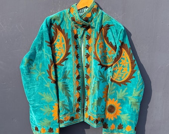 Op de afbeelding: Teal fluwelen jas met ingewikkelde borduursels in bruine en oranje bloempatronen. De jas heeft een hoge kraag, een ritssluiting aan de voorkant en gedetailleerde ontwerpen langs de mouwen en zoom. Opgehangen aan een houten hanger.