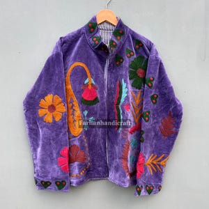 May include: A purple velvet jacket with colourful embroidered floral and paisley designs. The jacket features a zip-up front and a striped collar. The embroidery includes orange, green, pink, and blue accents. The jacket is hanging on a wooden hanger.