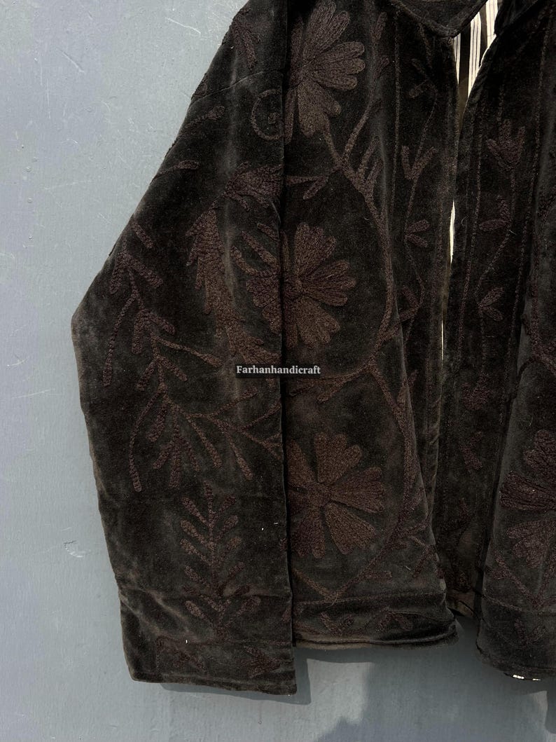 Op de afbeelding: Close-up van een donkerbruine fluwelen jas met bloemenborduurwerk. De jas heeft een gedetailleerd patroon van bloemen en bladeren. De tekst "Farhanhandicraft" is zichtbaar.