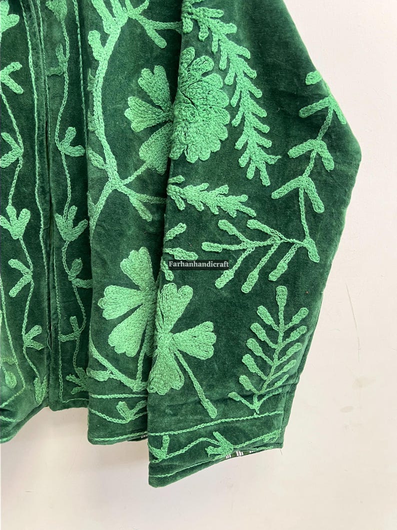 May include: Close-up of a dark green jacket with intricate light green floral embroidery. The jacket's fabric appears to be velvet, with detailed stitching of leaves and flowers. The text "Farhanhandicraft" is visible.