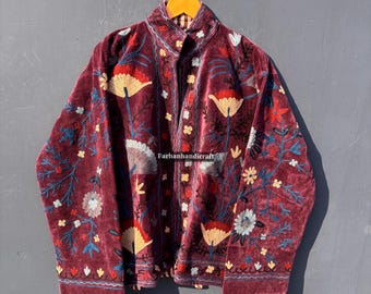 May include: Burgundy velvet jacket with floral embroidery in yellow, red, blue, and white. The jacket has a high collar and is buttoned up. The embroidery features stylized flowers and leaves. The jacket is hanging on a wooden hanger.
