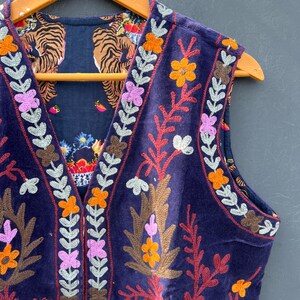 Puede incluir: Un chaleco sin mangas de terciopelo azul oscuro con bordados florales coloridos y un forro estampado con tigres. El chaleco est&aacute; adornado con dise&ntilde;os florales naranjas, rosas y blancos y se exhibe en una percha de madera.