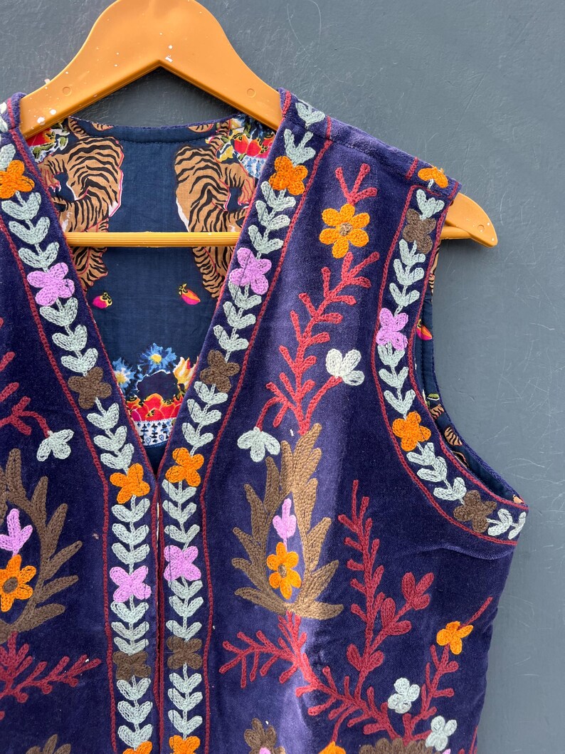 Puede incluir: Un chaleco sin mangas de terciopelo azul oscuro con bordados florales coloridos y un forro estampado con tigres. El chaleco est&aacute; adornado con dise&ntilde;os florales naranjas, rosas y blancos y se exhibe en una percha de madera.