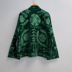 May include: A dark green velvet jacket with intricate floral embroidery in a lighter shade of green. The jacket is hanging on a wooden hanger against a plain white wall. The text "Farhanhandicraft" is visible.
