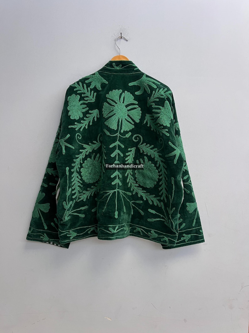 May include: A dark green velvet jacket with intricate floral embroidery in a lighter shade of green. The jacket is hanging on a wooden hanger against a plain white wall. The text "Farhanhandicraft" is visible.