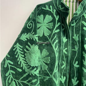 May include: A dark green velvet jacket with intricate light green floral embroidery. The jacket features a high collar and a striped lining. The embroidery includes flowers, vines, and leaves, creating a detailed pattern. The jacket is hanging on a wooden hanger.