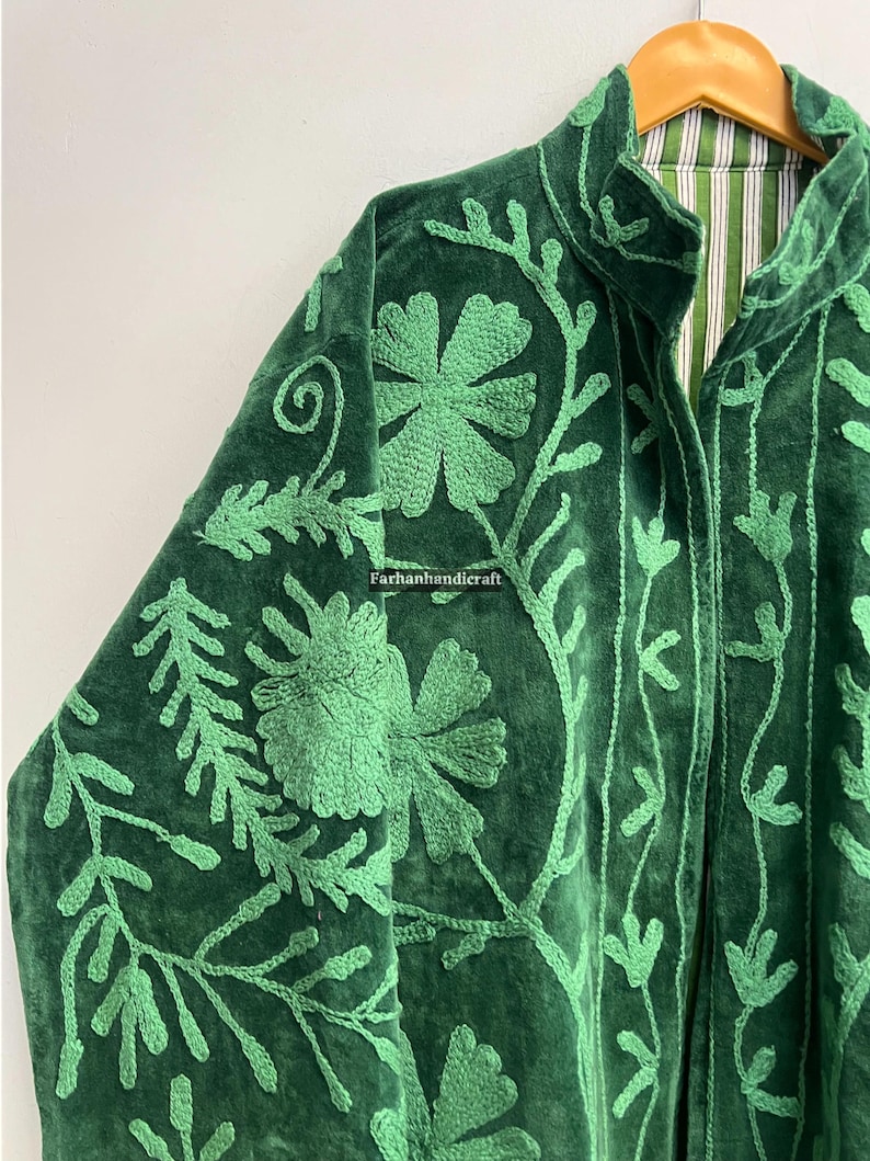 May include: A dark green velvet jacket with intricate light green floral embroidery. The jacket features a high collar and a striped lining. The embroidery includes flowers, vines, and leaves, creating a detailed pattern. The jacket is hanging on a wooden hanger.
