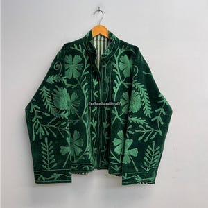 May include: A dark green velvet jacket with intricate floral embroidery. The jacket features a high collar and a striped lining. The embroidery is in a lighter shade of green, creating a contrasting design. The jacket is hanging on a wooden hanger.