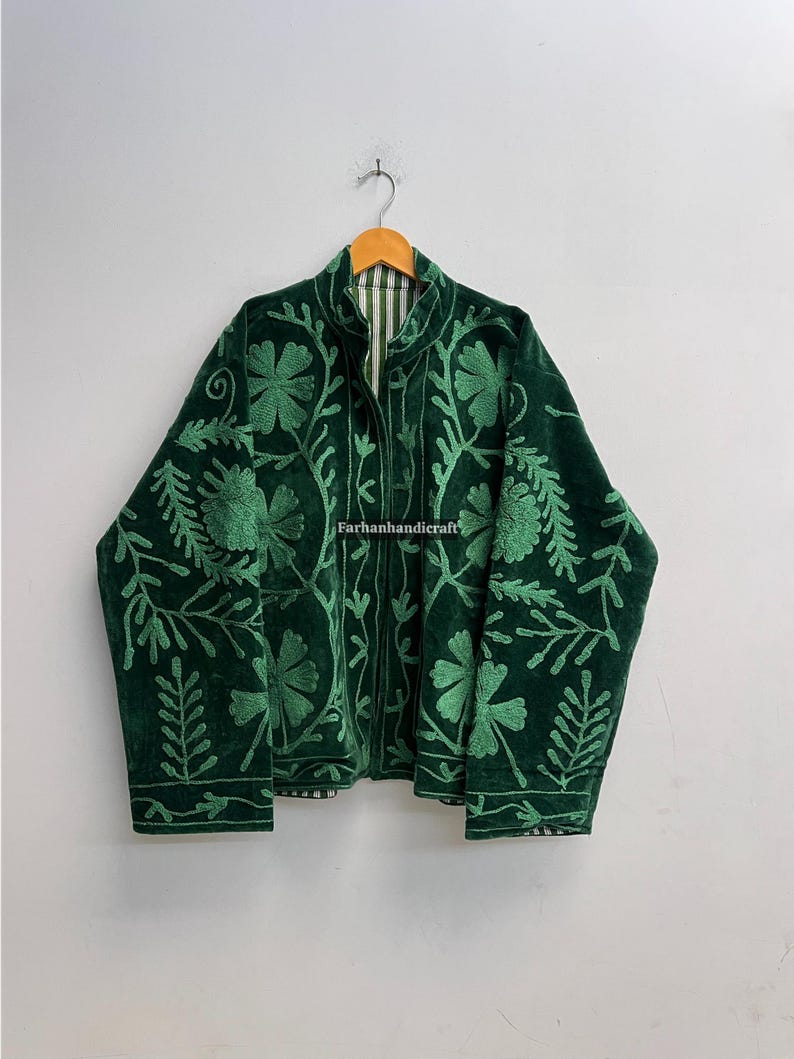 May include: A dark green velvet jacket with intricate floral embroidery. The jacket features a high collar and a striped lining. The embroidery is in a lighter shade of green, creating a contrasting design. The jacket is hanging on a wooden hanger.