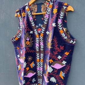 Puede incluir: Un chaleco de terciopelo azul marino con bordados florales coloridos. El chaleco presenta un cuello en V y es reversible, con un estampado de tigre en el interior. El bordado incluye flores naranjas, rosas y amarillas, y hojas marrones y verdes.