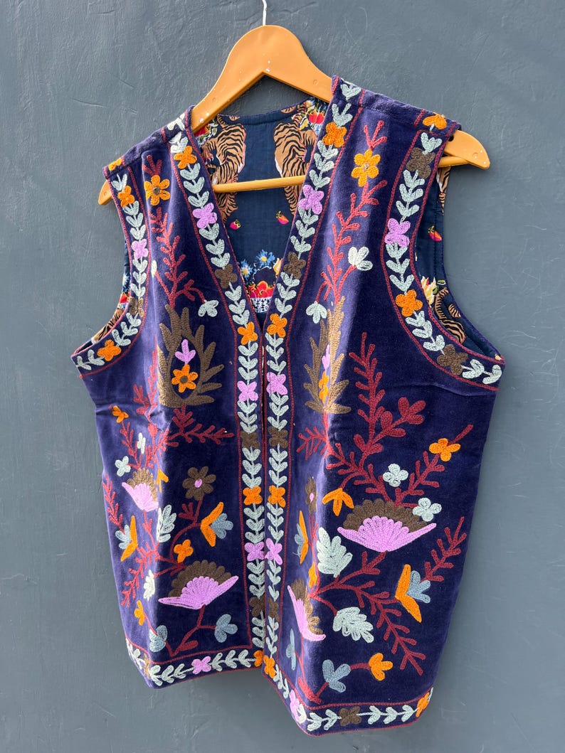 Puede incluir: Un chaleco de terciopelo azul marino con bordados florales coloridos. El chaleco presenta un cuello en V y es reversible, con un estampado de tigre en el interior. El bordado incluye flores naranjas, rosas y amarillas, y hojas marrones y verdes.