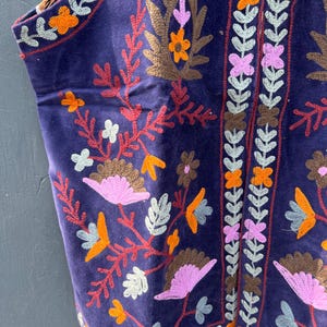 Puede incluir: Primer plano de un chaleco morado oscuro con intrincados bordados florales en tonos naranja, rosa, marr&oacute;n y blanco. El chaleco presenta un dise&ntilde;o detallado a lo largo de los bordes y un forro estampado contrastante.