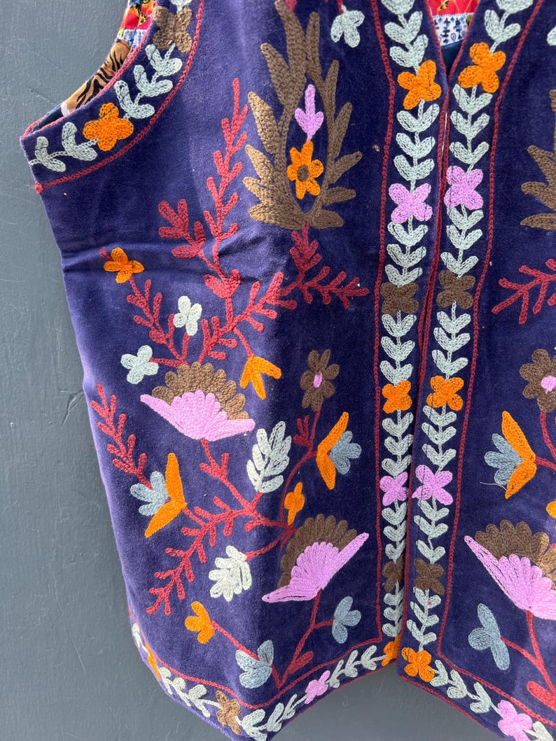 Puede incluir: Primer plano de un chaleco morado oscuro con intrincados bordados florales en tonos naranja, rosa, marr&oacute;n y blanco. El chaleco presenta un dise&ntilde;o detallado a lo largo de los bordes y un forro estampado contrastante.