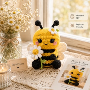 Puede incluir: Una abeja de ganchillo amarilla y negra con una margarita blanca, antenas negras y mejillas rosadas. La abeja se muestra junto a un folleto de patrón de ganchillo Print Velora, una tarjeta pequeña con el texto Print Velora y un florero.
