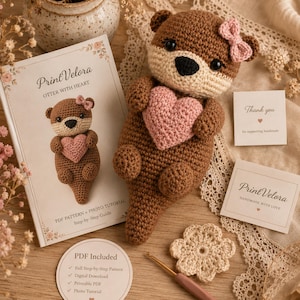 May include: A brown and cream crocheted otter plush holding a pink heart, accompanied by a PrintVelora instruction booklet, a thank you note, a small crochet flower, and a crochet hook. The booklet features the text PrintVelora Otter with Heart.