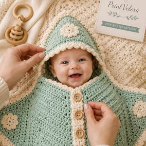 May include: A sage green crocheted baby blanket with a hood, featuring cream-colored scalloped edges, three wooden buttons down the front, and cream-colored floral appliques. The blanket is textured with a classic crochet stitch pattern.