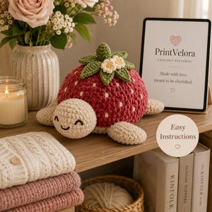 May include: A crocheted turtle toy with a red strawberry shell featuring white embroidered seeds and green leaves with two small white flowers. The turtle has a cream-colored head with a stitched smiling face and four matching cream-colored flippers.