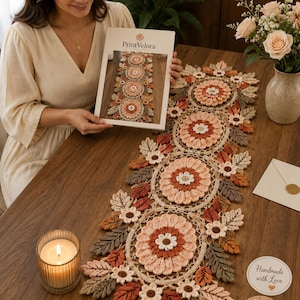 May include: A long, rectangular crochet table runner featuring a repeating pattern of large, circular floral motifs in shades of cream, peach, and rust, surrounded by intricate crocheted leaves in brown, tan, and orange, with a PrintVelora brochure.