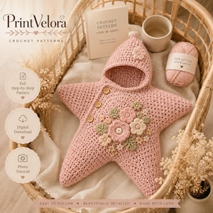 May include: A star-shaped pink crochet baby bunting with a hood, featuring three wooden buttons and a cluster of crocheted flowers and leaves in shades of pink, cream, and sage green. Includes a pattern booklet and a ball of pink cotton yarn.