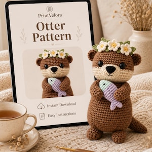 May include: A brown crocheted otter toy with a cream-colored face, wearing a green leaf and white flower crown, holding a light purple fish. Behind it, a tablet displays the text PrintVelora Otter Pattern, Instant Download, and Easy Instructions.