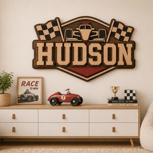 May include: A layered wooden wall sign featuring the name HUDSON in bold letters, centred below a race car graphic flanked by two chequered flags. The sign uses dark brown, light tan, and red wood tones to create a dimensional racing theme.