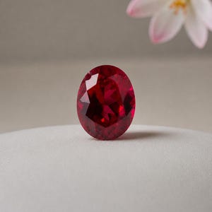 Puede incluir: Una gema de rubí facetada, de forma ovalada. La gema es de un color rojo intenso y rico, con reflejos internos visibles. El rubí está sobre una superficie blanca, con una flor borrosa en el fondo.