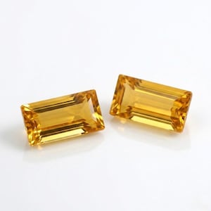 Peut inclure: Deux pierres précieuses rectangulaires jaune doré, taillées en brillant. Les gemmes sont posées sur un fond blanc, mettant en valeur leur clarté et leur couleur. Les pierres sont probablement de la citrine, une pierre précieuse populaire en joaillerie.