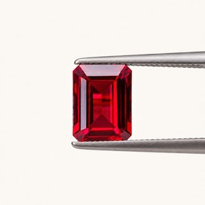 May include: A vibrant, deep red gemstone with an emerald cut, featuring sharp rectangular facets and a clear, transparent quality. The gemstone is held securely between the two metal tips of a pair of silver-toned tweezers.