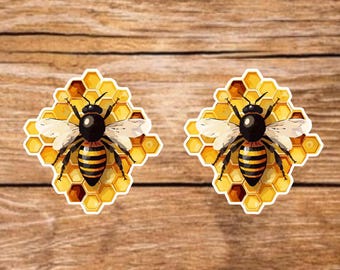 May include: Two bee stickers. Each sticker shows a black and yellow bee with white wings, set against a yellow honeycomb background. The stickers are on a wooden surface.
