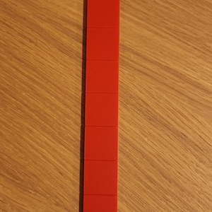 Op de afbeelding: Een verticale stapel van negen felrode, vierkante blokken. De blokken zijn gelijkmatig uitgelijnd en vormen een rechthoekige vorm. De blokken liggen op een houten oppervlak.