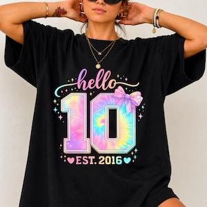 Puede incluir: Camiseta negra con un gráfico "10" tie-dye colorido, un lazo pastel y las palabras "hello" y "EST. 2016". El diseño incluye estrellas y corazones. La camiseta es de estilo casual.