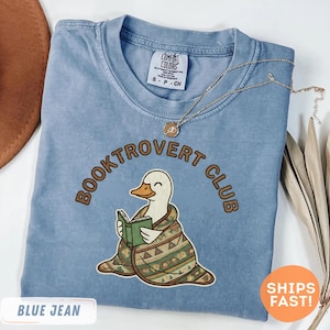 May include: A blue jean Comfort Colors t-shirt with the words "BOOKTROVERT CLUB" in brown lettering. The shirt features an embroidered duck reading a book, wrapped in a blanket. A gold necklace is draped across the shirt.
