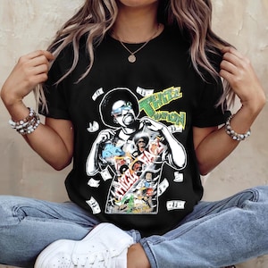 May include: A black short-sleeve t-shirt featuring a graphic print of a man with an afro wearing sunglasses. The design includes the text "THIZZ NATION" and "THIZZ FACE" in colorful lettering, surrounded by floating dollar bill illustrations.