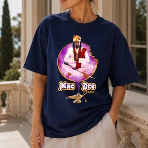 May include: A navy blue short-sleeve t-shirt featuring a graphic of a person in a white outfit and turban sitting cross-legged inside a purple circular cookie shape. Below, a gold genie lamp emits purple smoke with the text Mac Dre Cookies.
