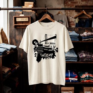 May include: A cream-colored short-sleeve t-shirt featuring a black graphic print. The design includes a person with an afro, a car, a bridge, mushrooms, and the text "THIZZ! R.I.P." along with the years "1970-2004" printed on the front center.
