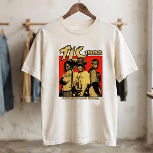 May include: A cream-coloured short-sleeve t-shirt featuring a graphic print of the music group TLC. The design includes the text TLC 1992 above a red square containing three figures and the text Ain't 2 Proud 2 Beg printed in gold lettering below.