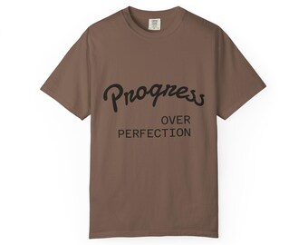 May include: A brown t-shirt featuring the black text "Progress Over Perfection". The shirt has a crew neck and short sleeves. The text is in a handwritten style font.