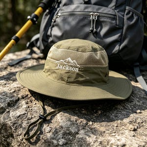 Peut inclure: Un chapeau bob vert olive doté d'une section de couronne en maille, d'un large bord et d'une mentonnière réglable. L'avant du chapeau présente un motif de montagne blanc brodé au-dessus du texte Jackson dans une police blanche sans empattement.