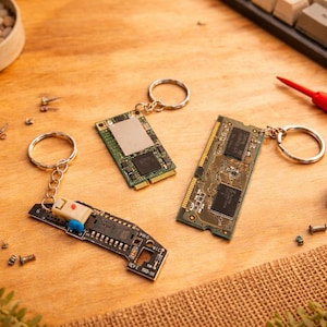 Könnte beinhalten: Drei Schlüsselanhänger mit Leiterplatten und silbernen Ringen werden auf einer Holzoberfläche präsentiert. Eine Spule aus Silberdraht, eine Sukkulente in einem kleinen Topf und eine Tastatur sind ebenfalls sichtbar. Kleine elektronische Bauteile sind verstreut.