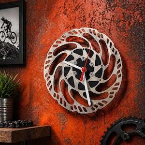 Könnte beinhalten: Wanduhr aus einer Fahrradbremsscheibe. Die Uhr hat einen silberfarbenen Rotor mit schwarzem Zentrum und weißen Zeigern mit rotem Sekundenzeiger. Sie ist an einer strukturierten orangefarbenen Wand befestigt.