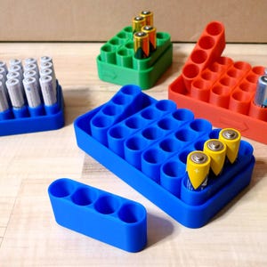 Könnte beinhalten: Eine Sammlung farbenfroher Batterie-Organizer in Blau, Grün und Rot. Die Organizer halten Batterien verschiedener Größen, einschließlich AA und AAA. Das Bild zeigt mehrere Organizer, die mit Batterien gefüllt sind und ihre Speicherkapazität und ihr Design zeigen.