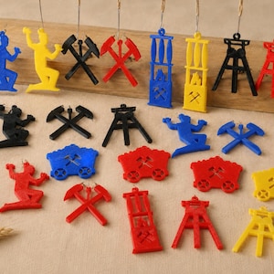 Könnte beinhalten: Eine Sammlung von hängenden Ornamenten und Anhängern in Rot, Blau, Gelb und Schwarz. Die Formen umfassen kniende Figuren, gekreuzte Hämmer, Förderwagen und Fördertürme. Jedes Teil verfügt über eine kleine Öse an der Oberseite zur Befestigung.