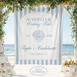 May include: A light blue and white vertical striped fabric banner featuring a central arch design with the text SEASHELLS & Wedding Bells, EST. 2026, Taylor's Bachelorette, and LAKE TAHOE, CA, centered around a detailed line drawing of a seashell.