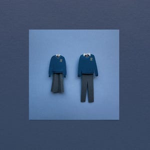 May include: Two paper cut-outs of school uniforms, one blue sweater with a skirt and the other a blue sweater with trousers, both with a crest on the front.