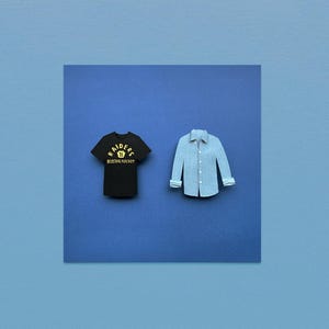 May include: A black t-shirt with the text "Raiders Boston Hockey" in gold lettering and a light blue button-down shirt. Both shirts are displayed on a dark blue square against a lighter blue background.