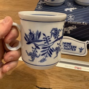 Puede incluir: Taza de cerámica blanca con un patrón floral azul y blanco. La taza tiene un asa y dice "Genuine Pattern Painted" en un pequeño letrero blanco.